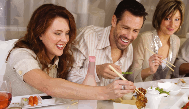 Three people eating with chopsticks, two of whom are laughing