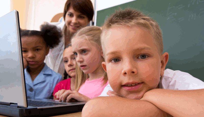 A child looks at the camera while his classmates study a laptop computer