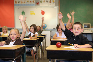 Children seated at desks in a classroom with their arms raised