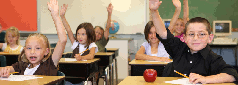 Children seated at desks in a classroom with their arms raised