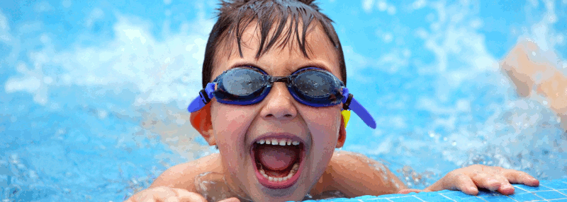 A young boy with swimming goggles grins over the edge of a pool